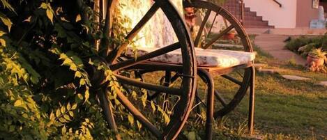 A rustic outdoor seating area with a metal bench and large wheels, surrounded by greenery and a staircase leading to a building.