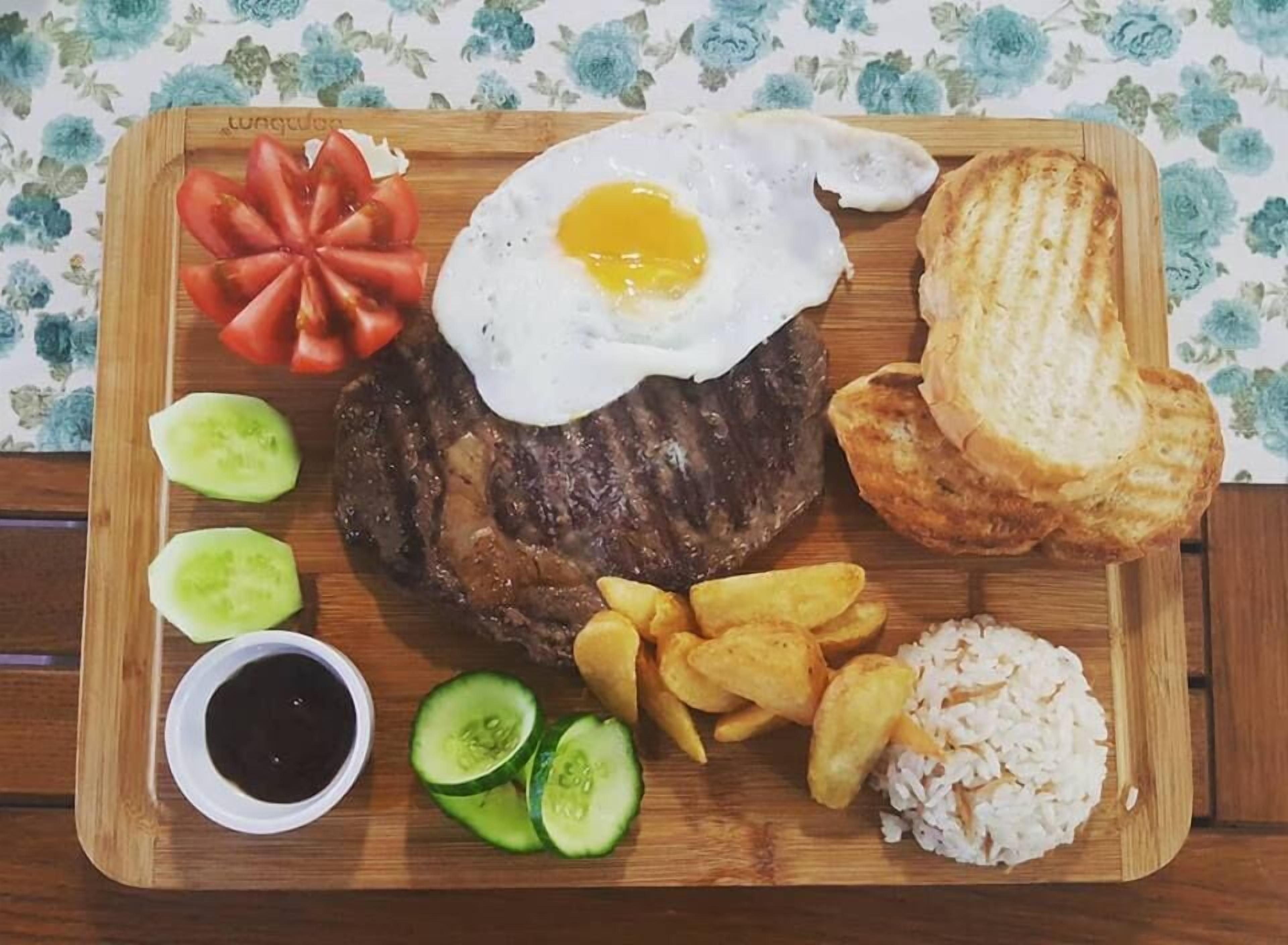 A wooden board with a steak, fried egg, toast, French fries, cucumber slices, and rice.