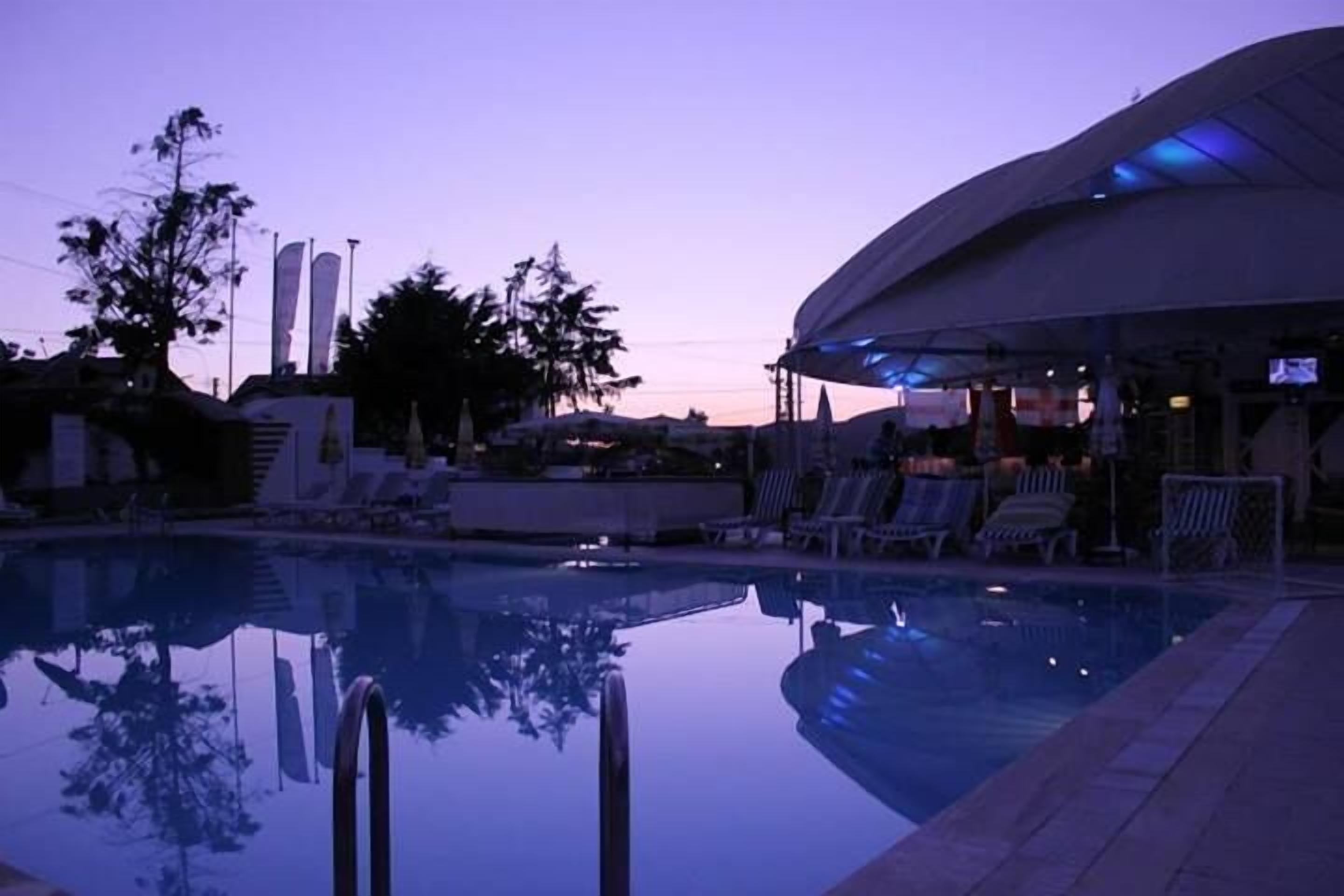 Una piscina con sillones de descanso y una zona cubierta, rodeada de árboles y edificios.