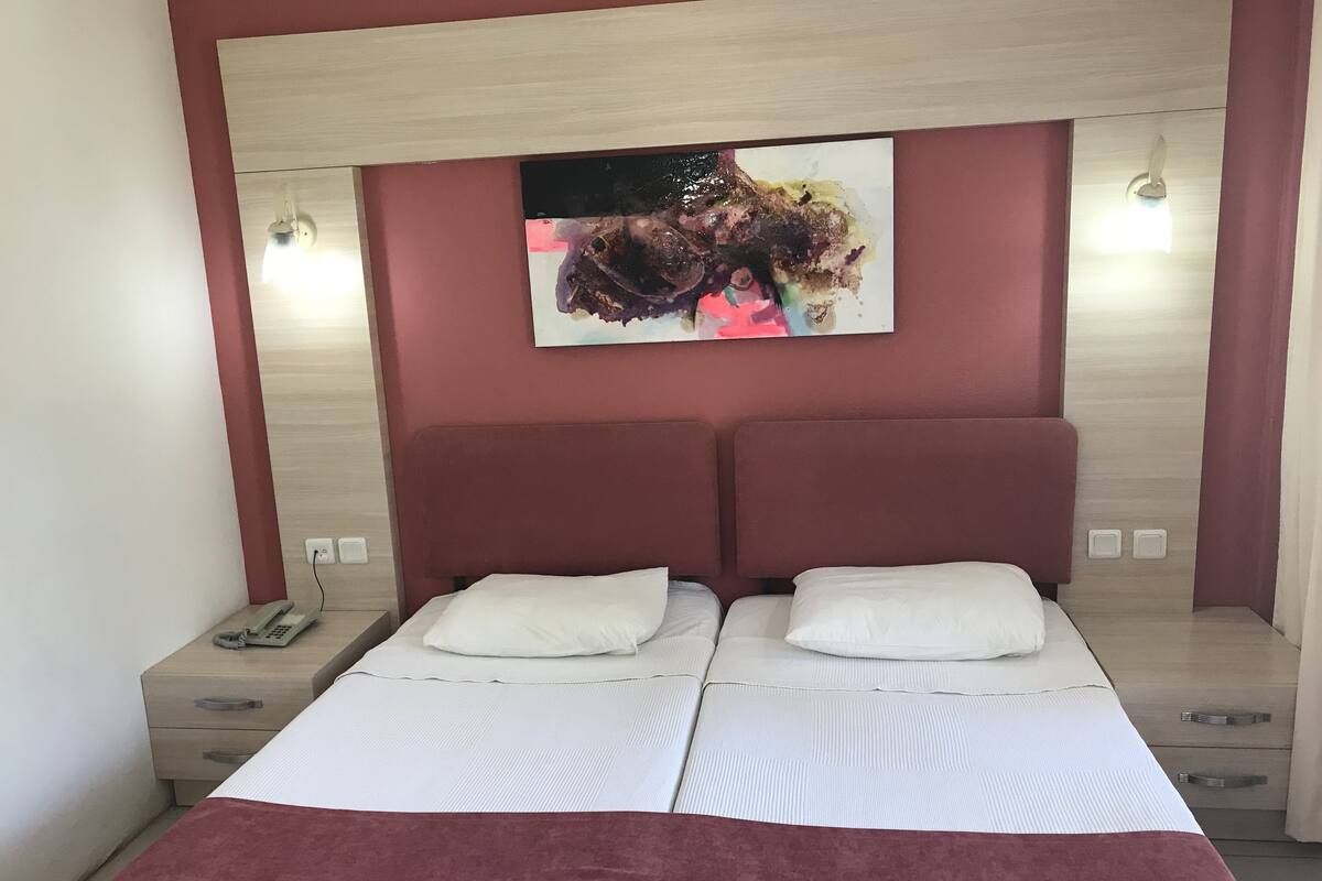 Uma cama de casal com roupa de cama branca e duas almofadas, cabeceira de madeira, um quadro acima da cama e luminárias de cabeceira.