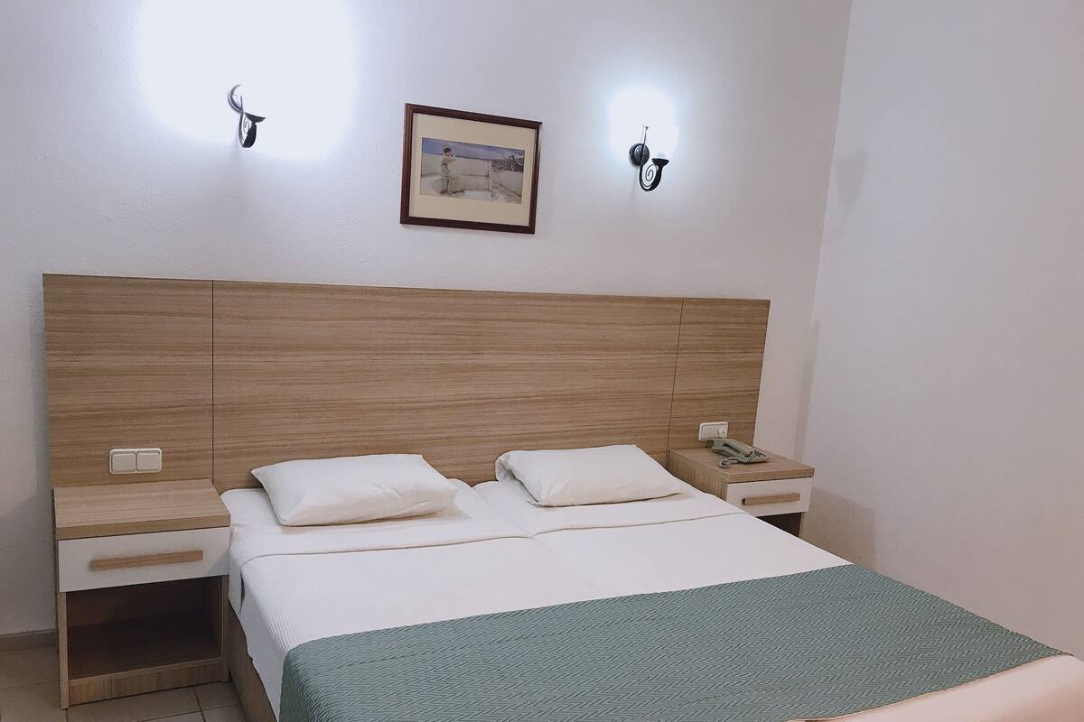 Uma cama bem arrumada com lençóis brancos e um manta verde, duas mesas de cabeceira com abajures e um quadro na parede.