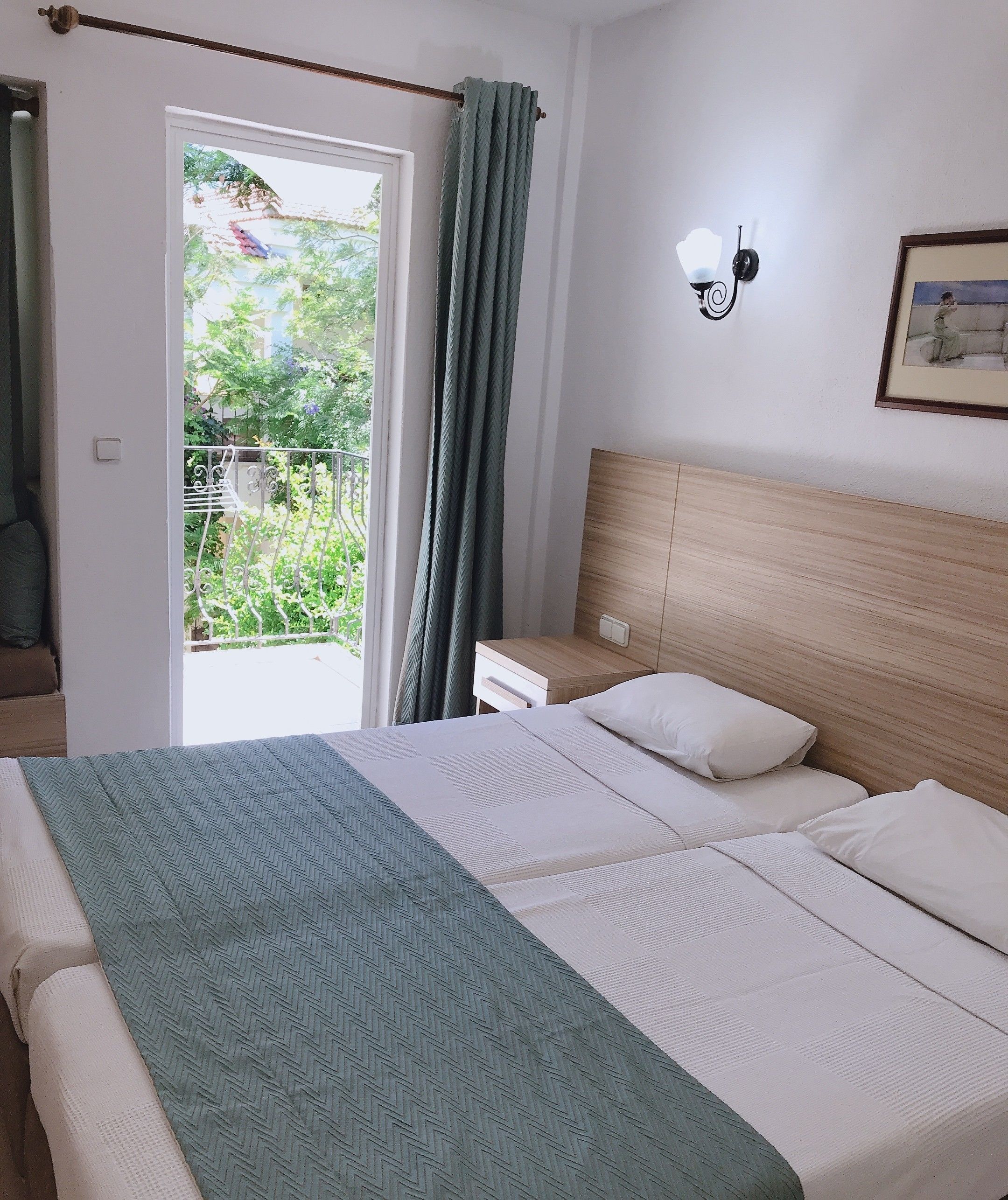 A bedroom with a bed, a nightstand, a wall-mounted light, and a view of greenery outside.