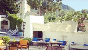 An outdoor seating area with wooden furniture and cushions, a pool in the background, and a mountainous landscape.