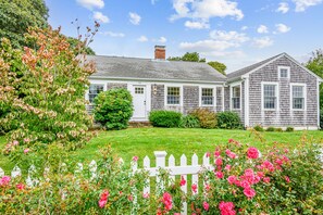 A two-story house with a white picket fence, a well-maintained lawn, and a variety of flowering plants. - Your Perfect South Chatham Summer Stay!! (Chatham)