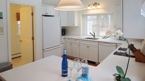 A modern kitchen with white cabinets, a central island with a bottle of wine and glasses, a ceiling fan, and a window with a view.