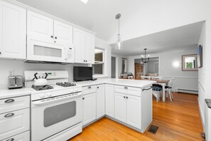A modern kitchen with white cabinetry, a gas stove, a microwave, and a dining area with a table and chairs. - Your Perfect South Chatham Summer Stay!! (Chatham)