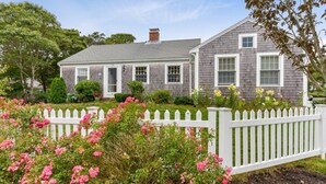 A two-story house with a white picket fence, surrounded by lush greenery and pink flowers.