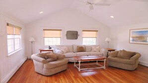 A spacious living room with a high ceiling, wooden flooring, and a ceiling fan. There are two windows with blinds, a painting on the wall, and a small table with a lamp.