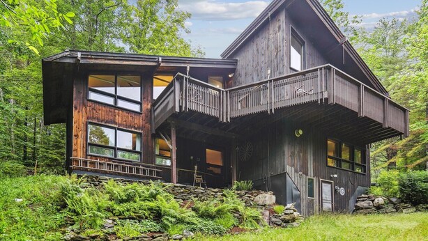 Eine rustikale Holzhütte mit großem Balkon, umgeben von üppigem Grün und einem Steinfundament.