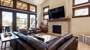 Un salón amplio con chimenea de piedra, sofás de cuero, televisor de pantalla plana y ventanas grandes que ofrecen vistas al exterior.