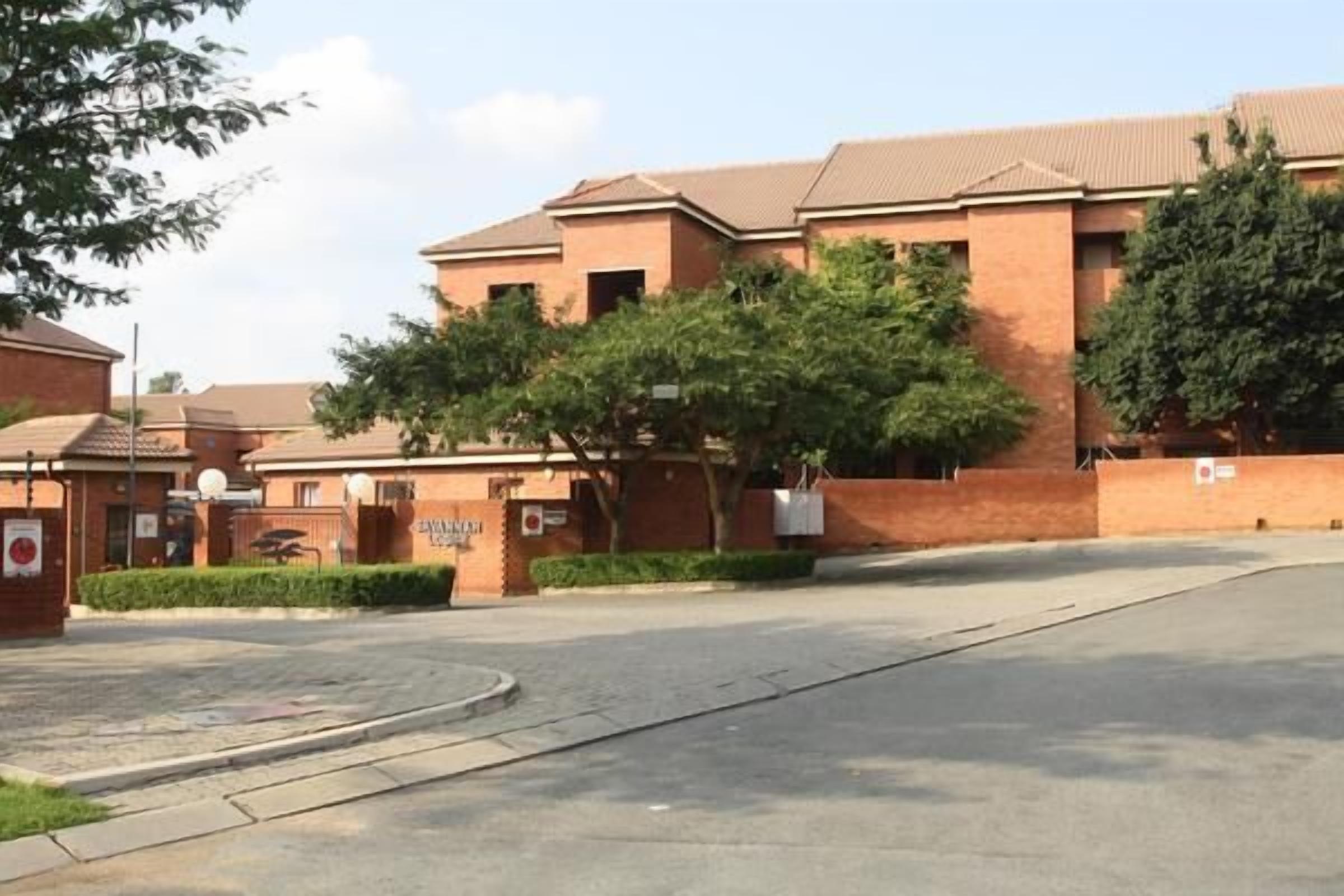 A brick building with a flat roof, surrounded by greenery and a paved area.