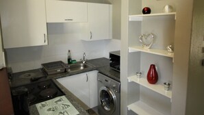 A compact kitchen with white cabinets, a built-in washing machine, a granite countertop, and a small shelf with decorative items.