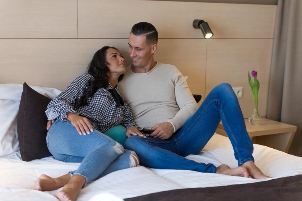 Una pareja sentada en una cama en una habitación de hotel, con una mesita de noche y un jarrón con una flor.