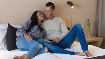 Una pareja sentada en una cama en una habitación de hotel, con una mesita de noche y un jarrón con una flor.