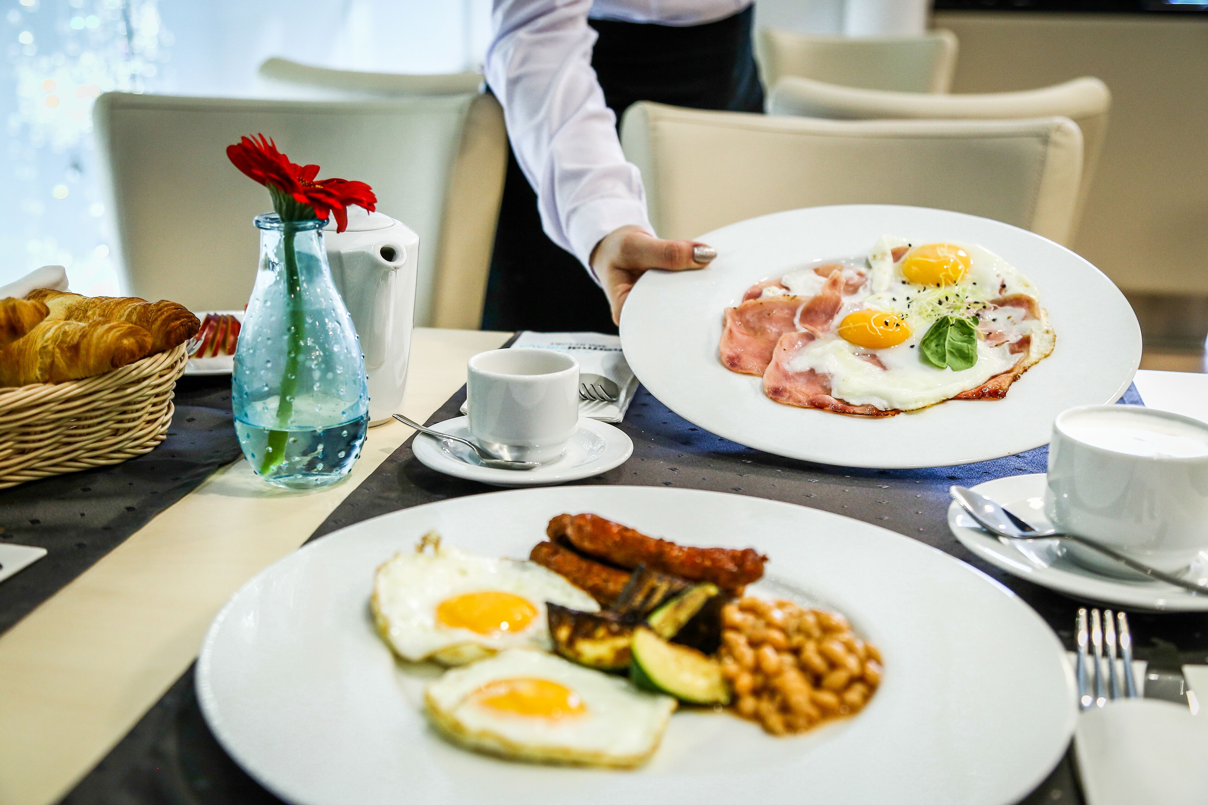A breakfast setting with plates of eggs, sausages, and toast, a bottle of water, and a basket of croissants.