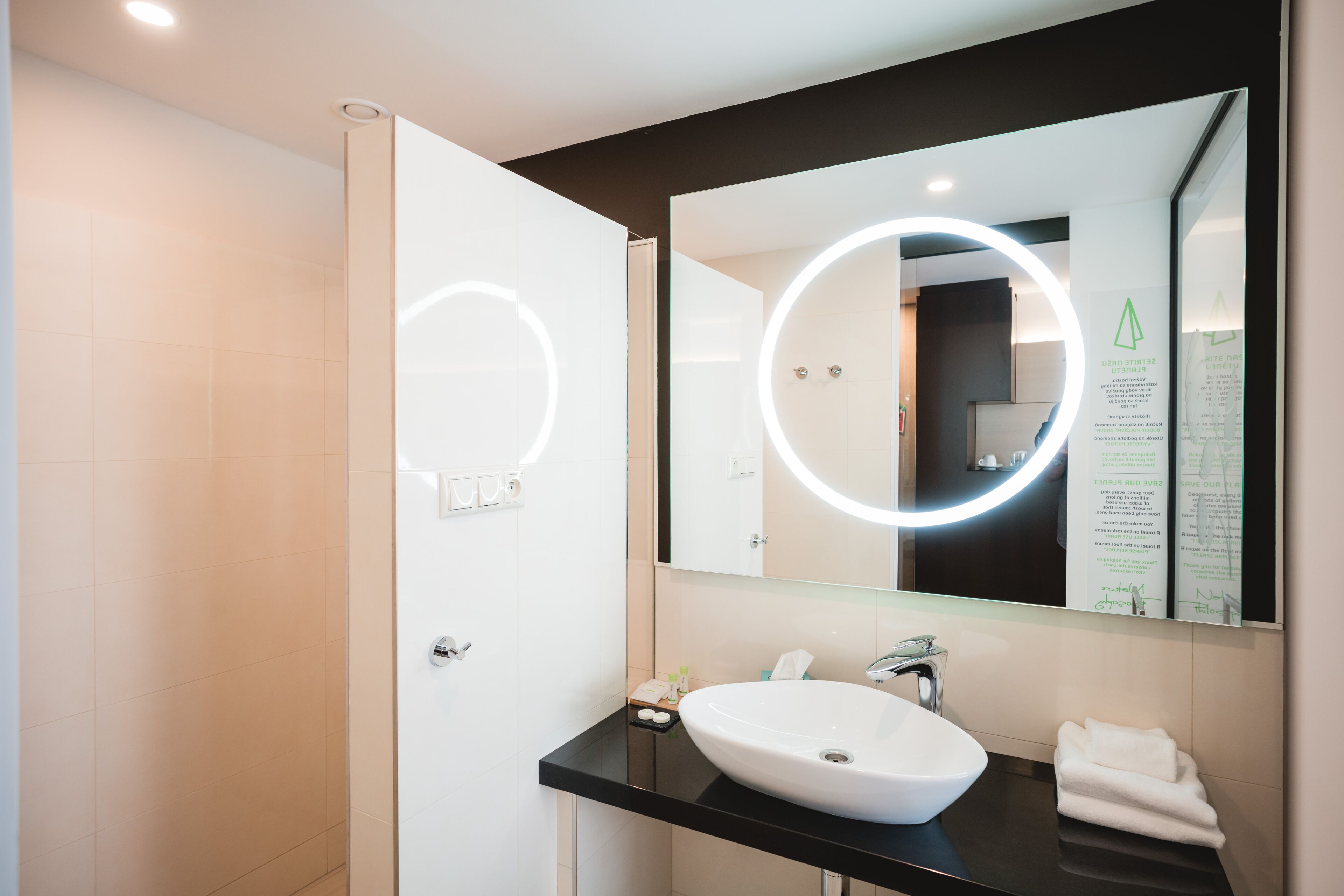 Casa de banho moderna com lavatório branco, bancada preta e um espelho grande com luz circular.