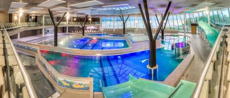 Una gran piscina cubierta con múltiples carriles y una barrera de cristal. Hay zonas de estar con sillas y mesas alrededor de la piscina.