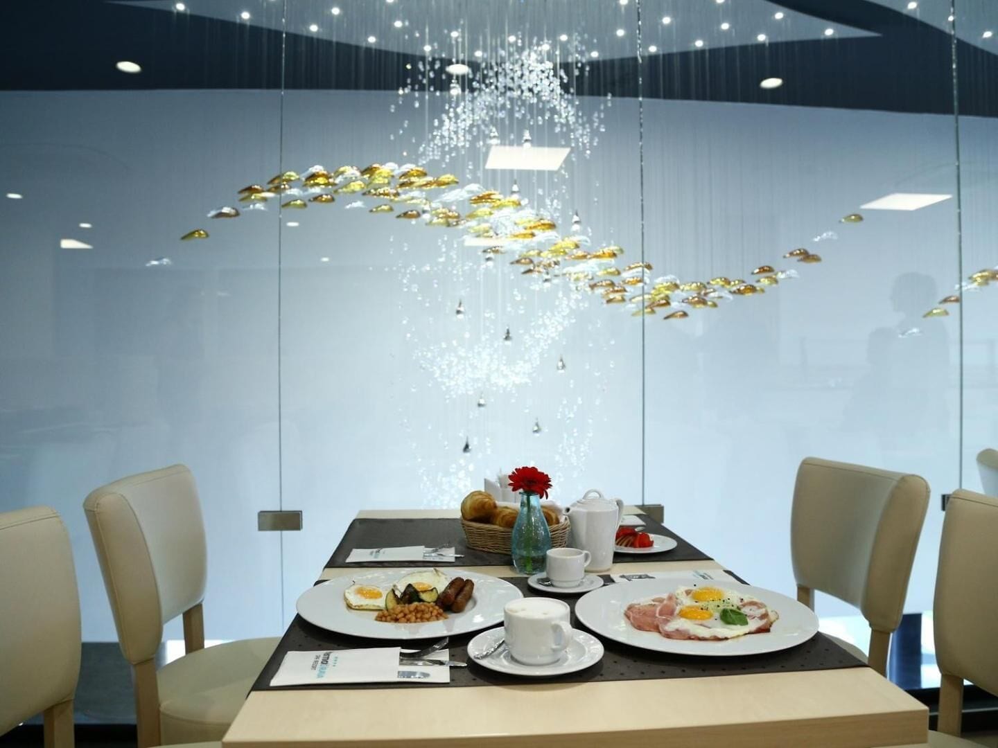Een eettafel met ontbijtbestek, een vaas met een rode bloem en een hanglamp.