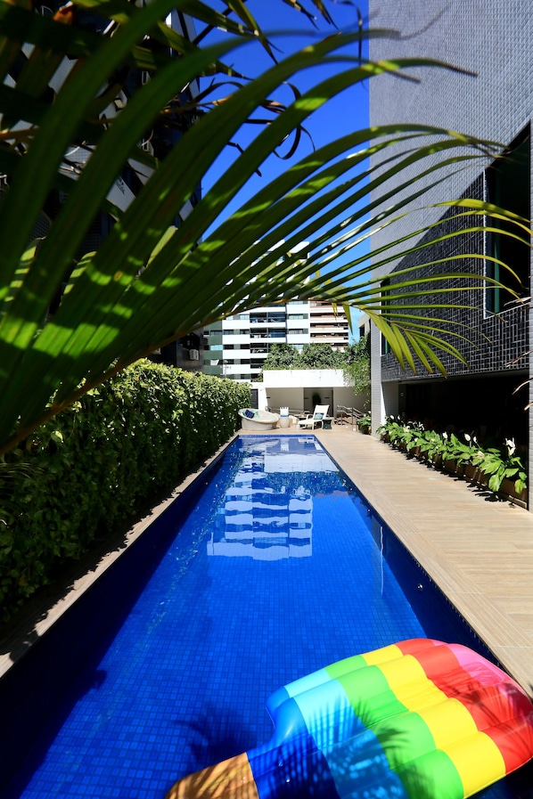 A swimming pool with a rainbow-colored inflatable float, surrounded by greenery and modern buildings.