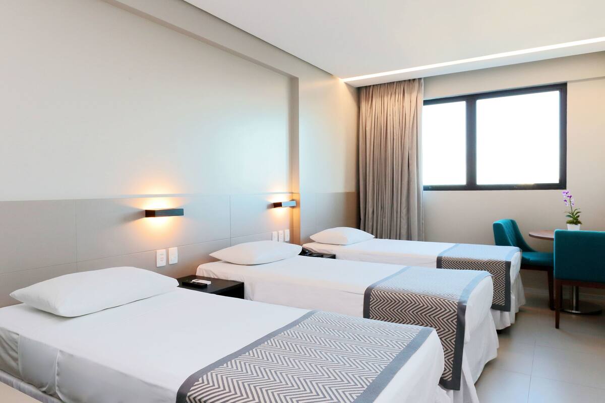 A hotel room with two single beds, a desk with a chair, a window with curtains, and wall-mounted lights.