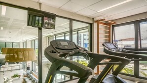 A modern gym with treadmills, a TV mounted on the wall, and a view of the outside through large windows. - Porto Kaeté Hotel (Maceió)