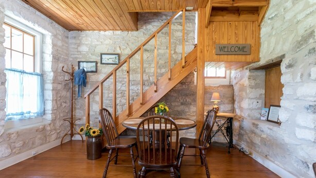Un interior rústico con una escalera de madera, paredes de piedra, una mesa redonda de comedor con sillas y un letrero que dice 'WELCOME'.