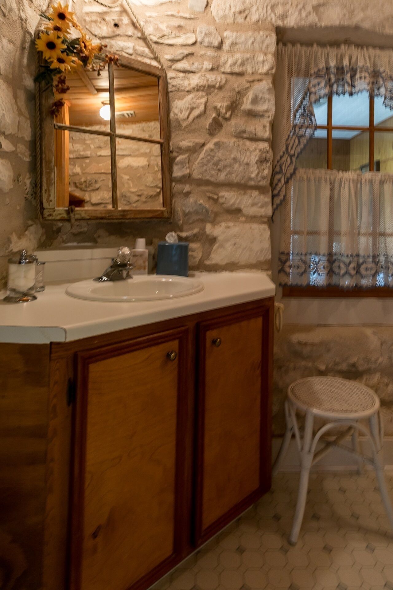 Une salle de bain avec un mur en pierre, des meubles en bois, un lavabo blanc et un tabouret en osier.