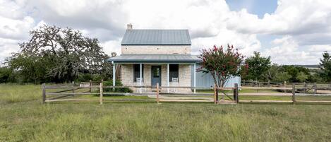 A stone building with a blue roof and a porch, surrounded by a wooden fence and a grassy field.