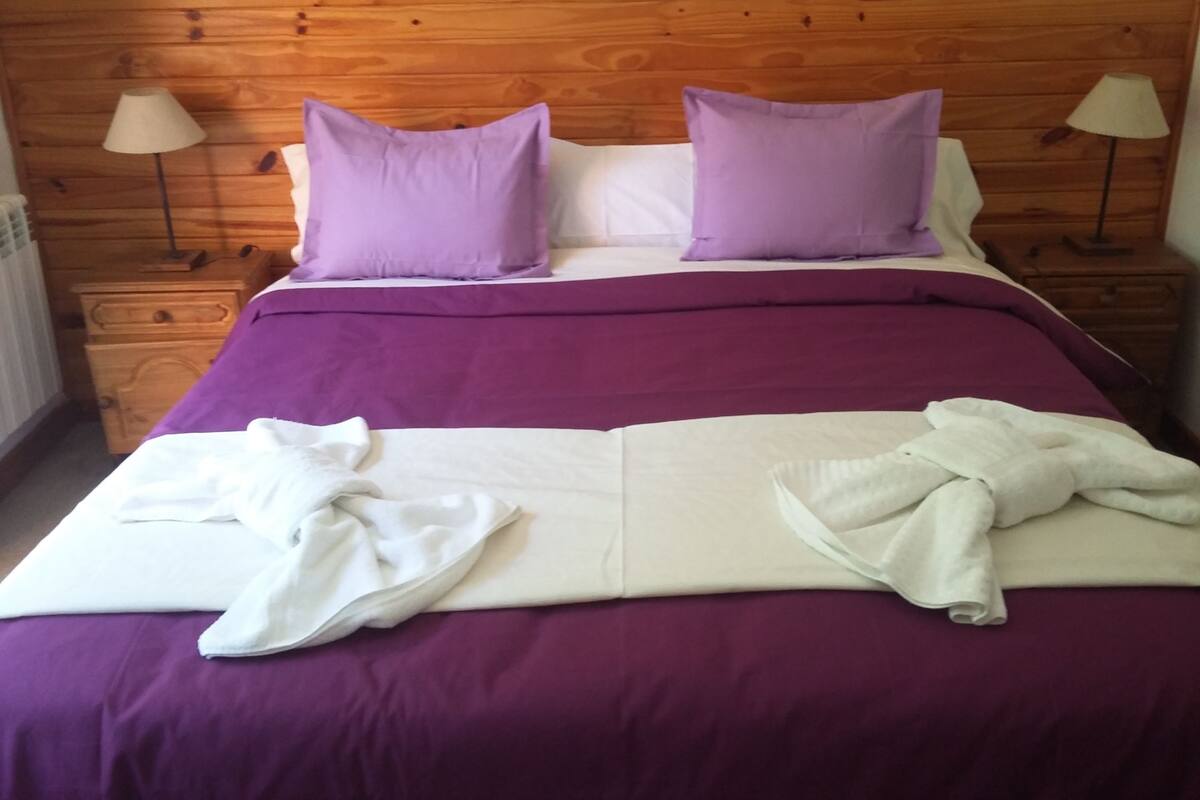 A neatly made bed with purple bedding and white towels, wooden headboard, and two bedside lamps.