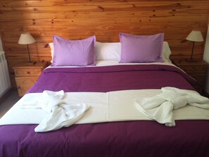 Ein ordentlich bezogenes Bett mit violetter Bettwäsche und weißen Handtüchern, ein hölzerner Kopfteil und zwei Nachttischlampen.