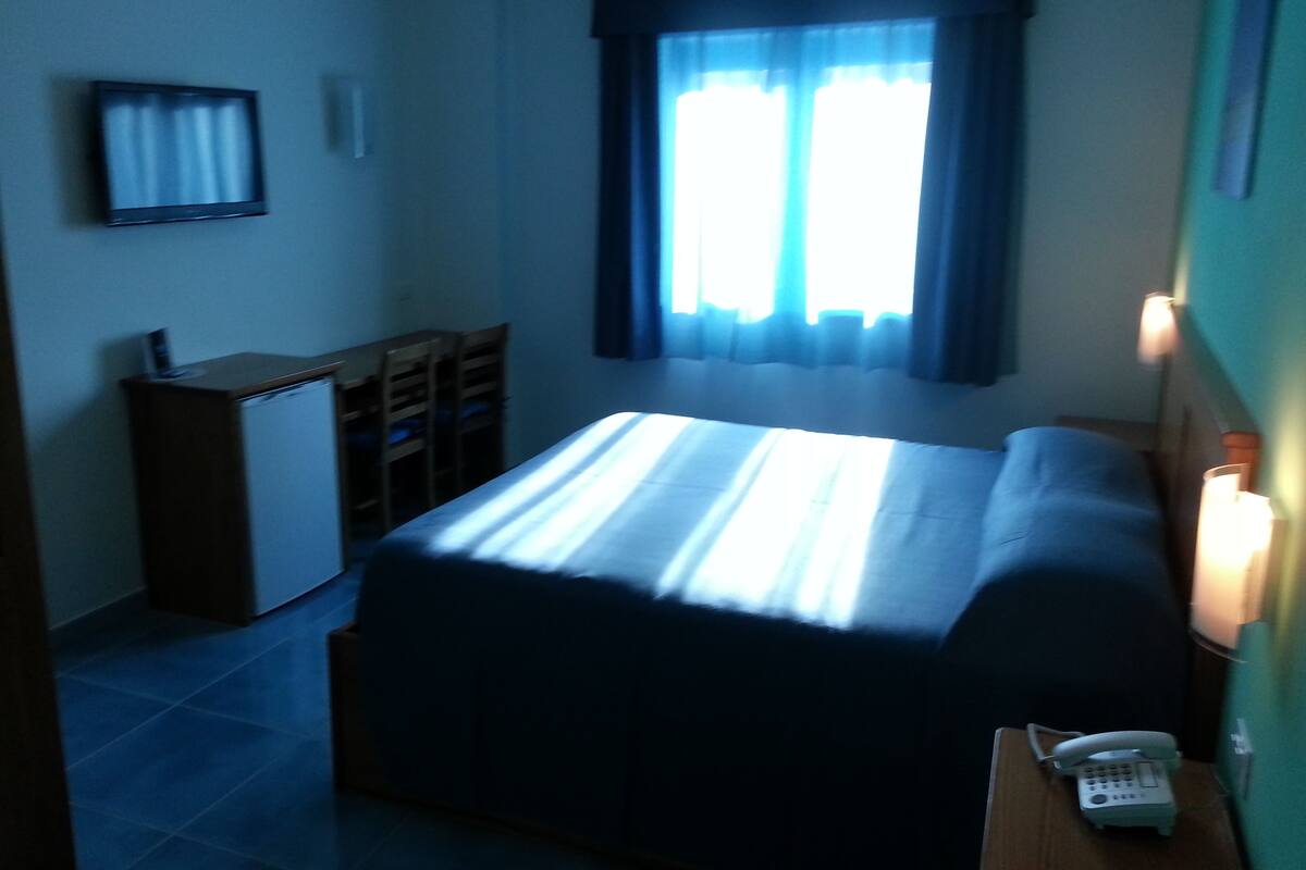 A small, dimly lit room with a bed, a desk, and a chair. There is a window with curtains, a wall-mounted TV, and a bedside table with a phone.