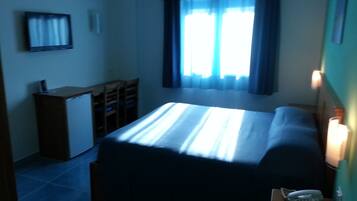 Ein kleiner, schummrig beleuchteter Raum mit einem Bett, einem Schreibtisch und einem Stuhl. Es gibt ein Fenster mit Vorhängen, einen an der Wand montierten Fernseher und einen Nachttisch mit einem Telefon.