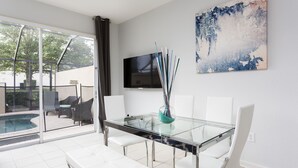 A modern dining area with a glass table, white chairs, a TV on the wall, and a view of a pool and outdoor seating.