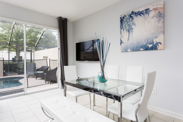 Un comedor moderno con una mesa de vidrio, sillas blancas, un televisor en la pared y vistas a una piscina y zona de estar al aire libre.