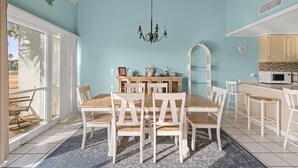 A dining area with a wooden table and white chairs, a chandelier, and a kitchen with a microwave.