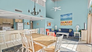 A spacious living area with a dining table, chairs, a sofa, and a coffee table. There is a kitchen with a refrigerator and cabinets. The walls are painted light blue, and there is a ceiling fan.
