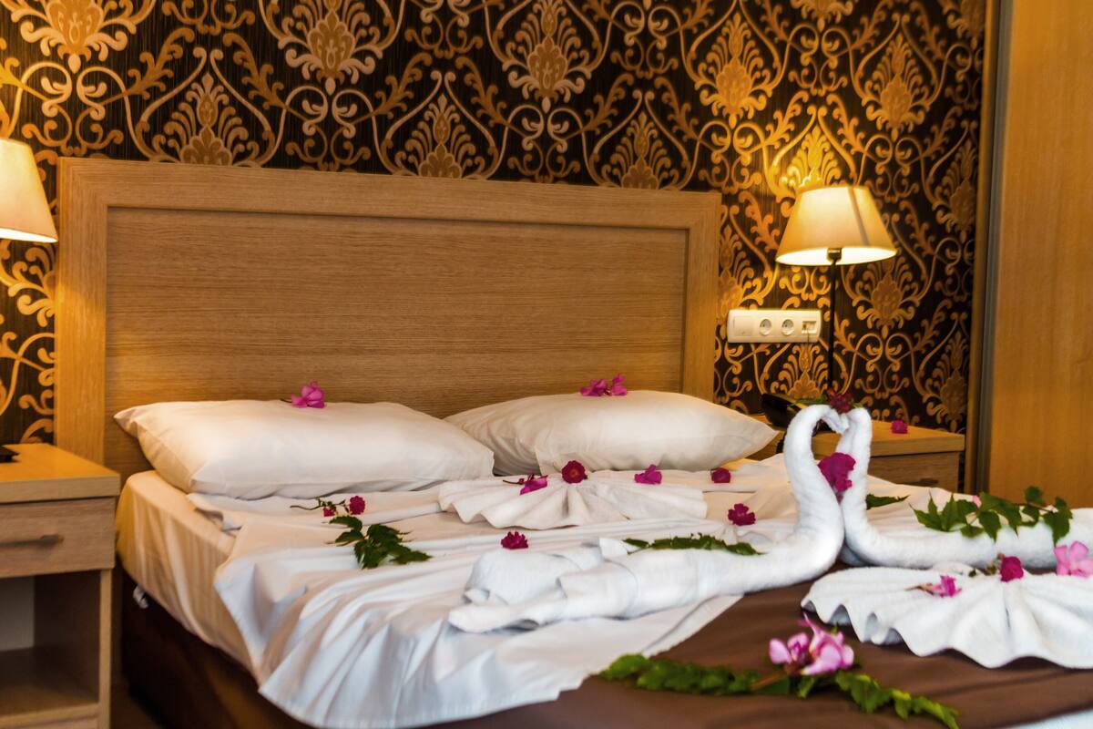 A neatly made bed with white linens, two pillows, and a swan-shaped towel arrangement. There are two bedside lamps and a wall with an ornate pattern.