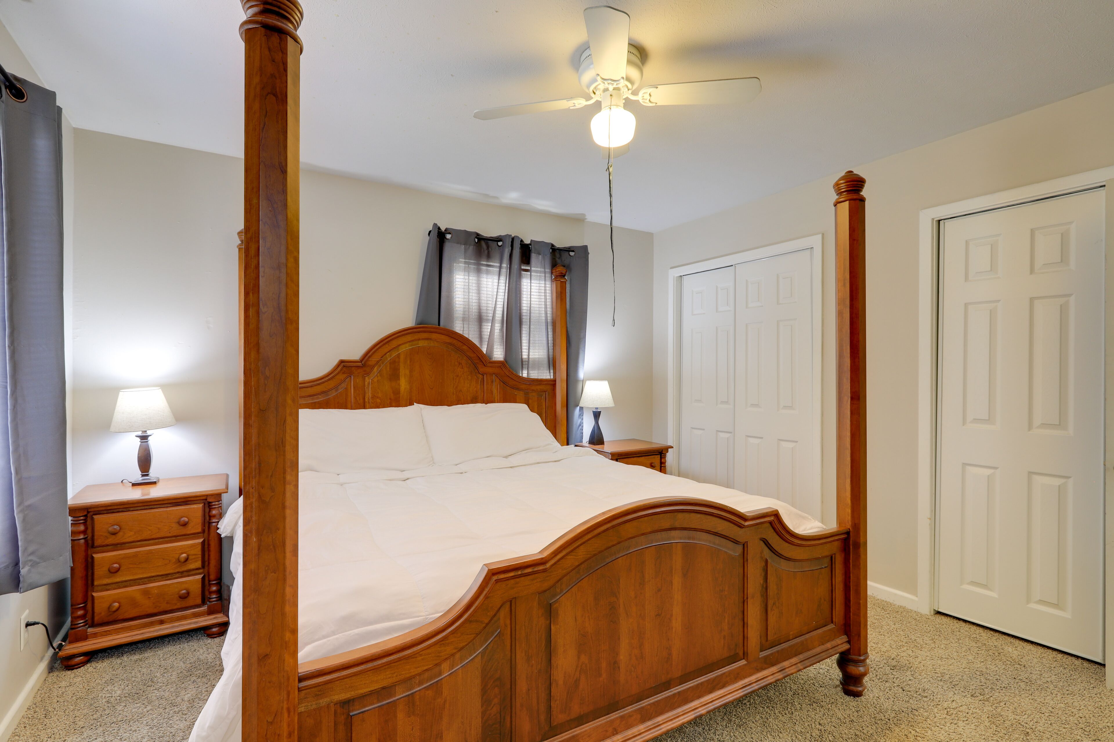 A four-poster bed with a wooden headboard and footboard, two bedside tables with lamps, a ceiling fan, and a white door.