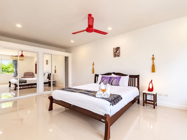 A spacious bedroom with a large bed, a red ceiling fan, and a view through a glass door to an outdoor area.
