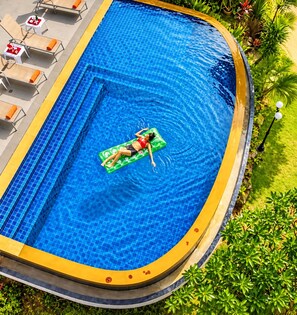 Piscina com uma pessoa boiando, cercada por espreguiçadeiras e um jardim bem cuidado.