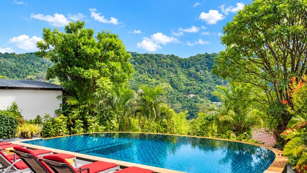 A swimming pool with red lounge chairs, surrounded by lush greenery and a mountainous backdrop. - Villa Nap Dau Crown Private pool villa for Family and Friends (ต.ฉลอง)