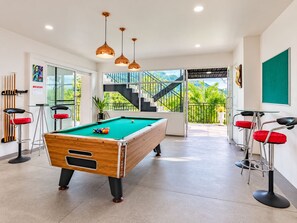 A pool table in a room with a staircase leading to a balcony, a green chalkboard, and two bar stools. - Villa Nap Dau Crown Private pool villa for Family and Friends (ต.ฉลอง)