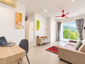 Una sala de estar moderna con una computadora portátil sobre un escritorio, un sofá, un ventilador de techo rojo y vistas a la vegetación a través de la ventana.