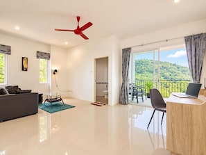 A modern living room with a sofa, a dining table with chairs, a red ceiling fan, and a balcony with a view of a forested hill.