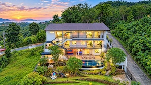 Hotel de varios pisos con piscina, rodeado de exuberante vegetación y vista al atardecer.