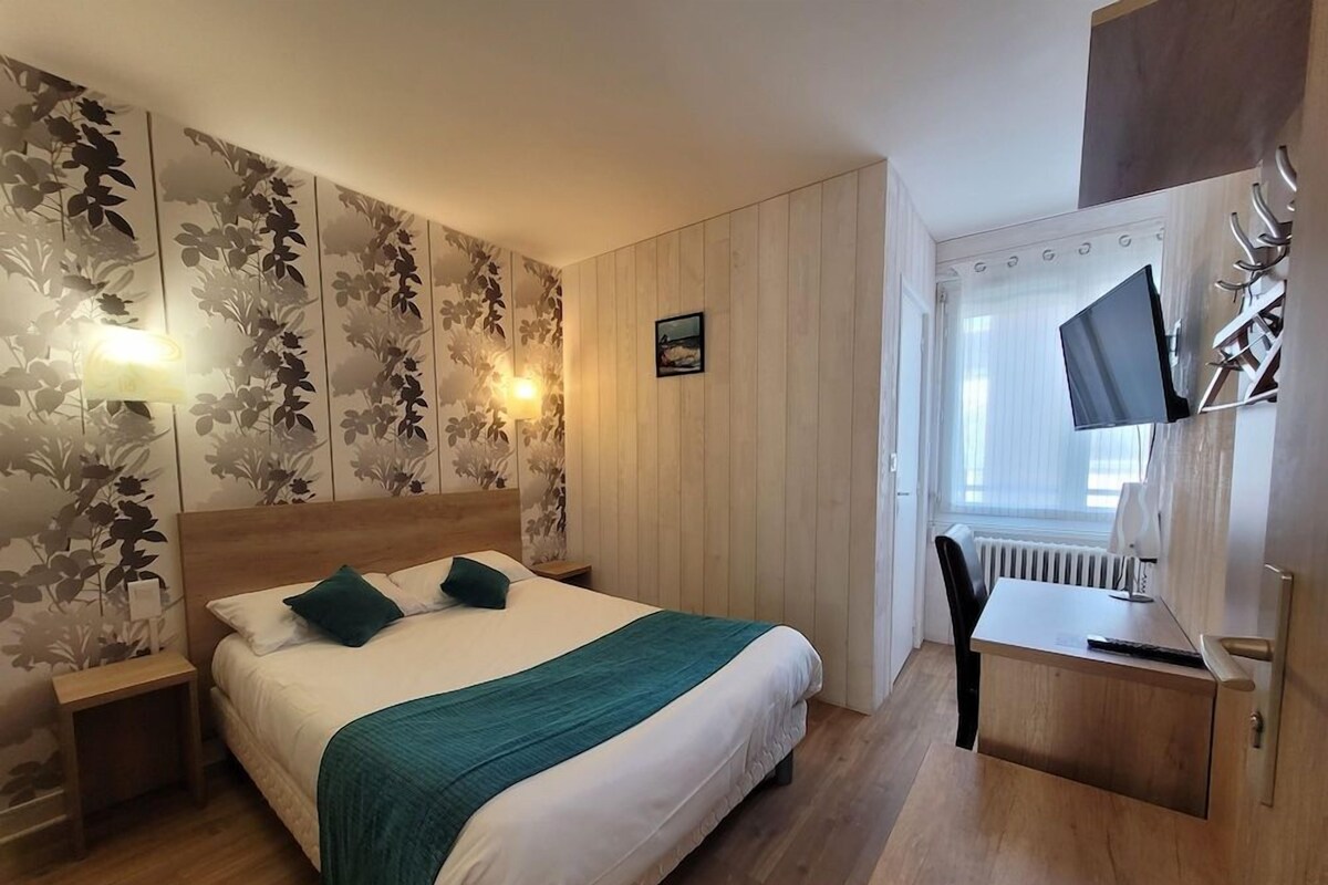 A hotel room with a bed, bedside table, lamp, TV, and a desk with a chair.