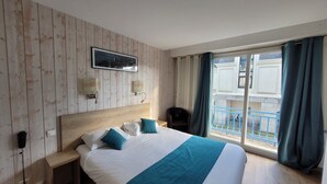 Habitación de hotel con pared de paneles de madera, cama con ropa de cama blanca y turquesa, mesita de noche pequeña, una silla y una ventana con cortinas.