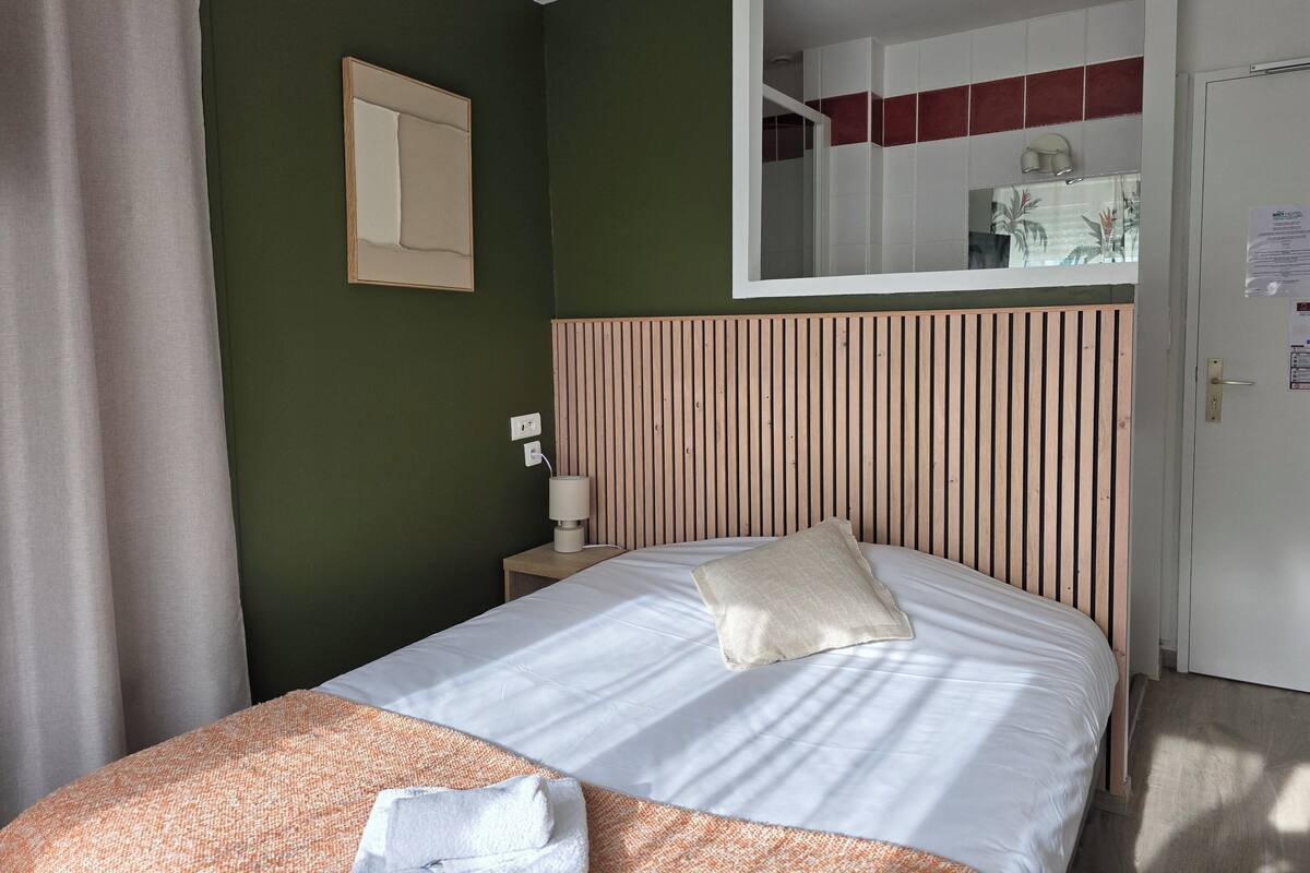 A neatly made bed with a pillow and a folded towel, a bedside lamp, a mirror, and a door with a red stripe.