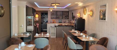 Un moderno comedor con mesas y sillas de madera, una cocina al fondo e iluminación colgante.