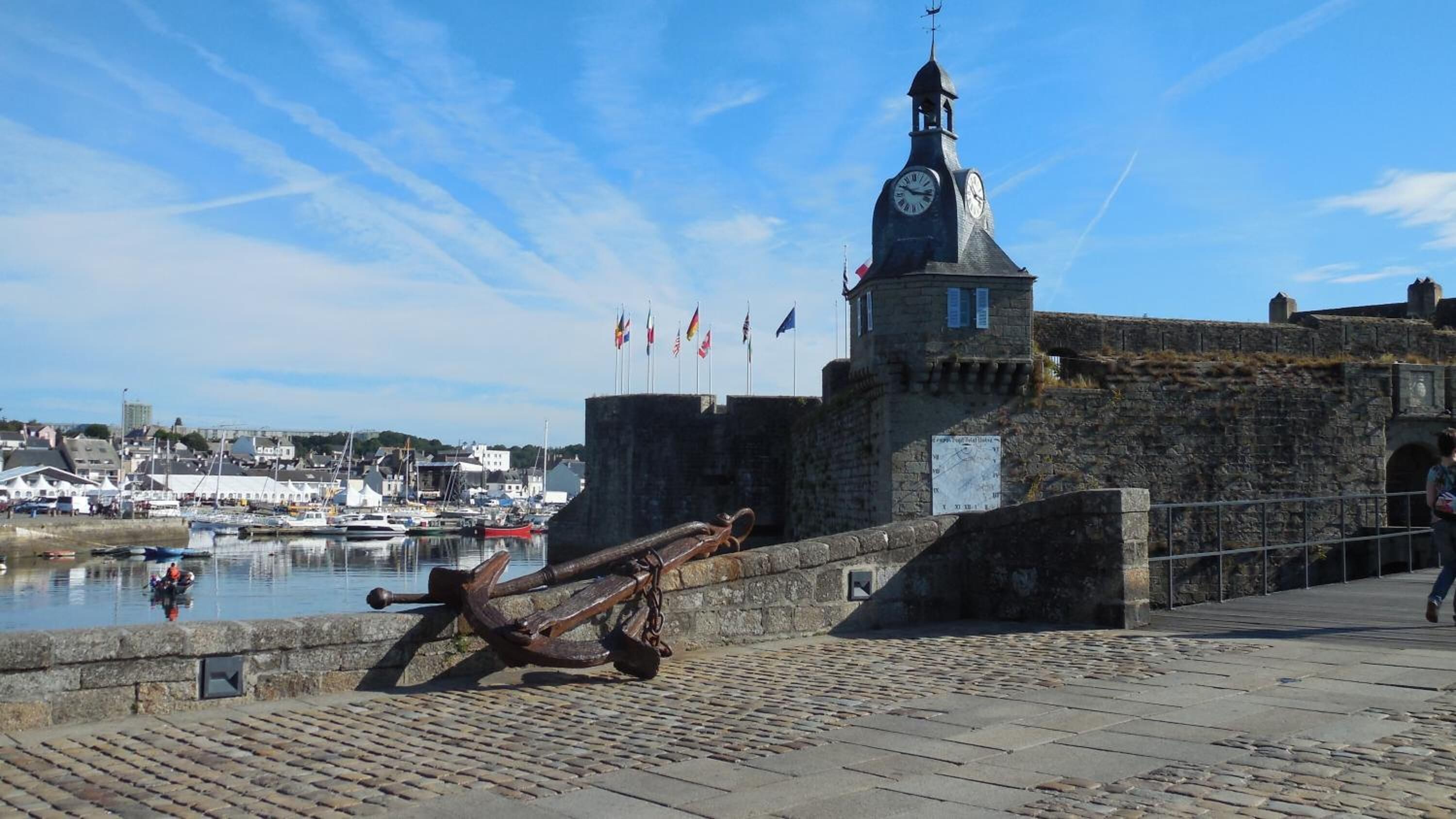 Un edificio histórico de piedra con una torre del reloj, vistas al agua y una gran escultura de ancla.